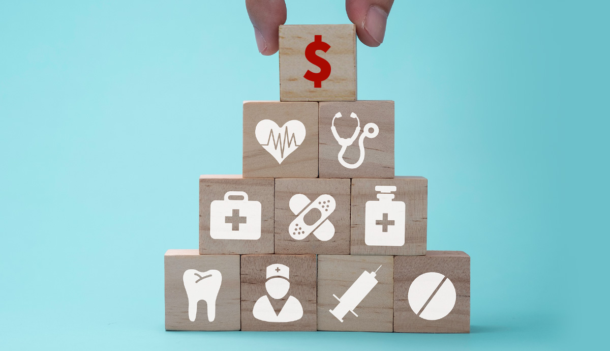 A hand places a building block featuring a red dollar sign on top of stacked blocks featuring assorted healthcare-related images.