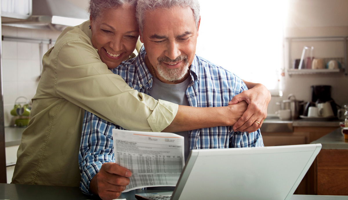 A smiling woman with her arms around a smiling man looking at a bill together in front of a laptop.