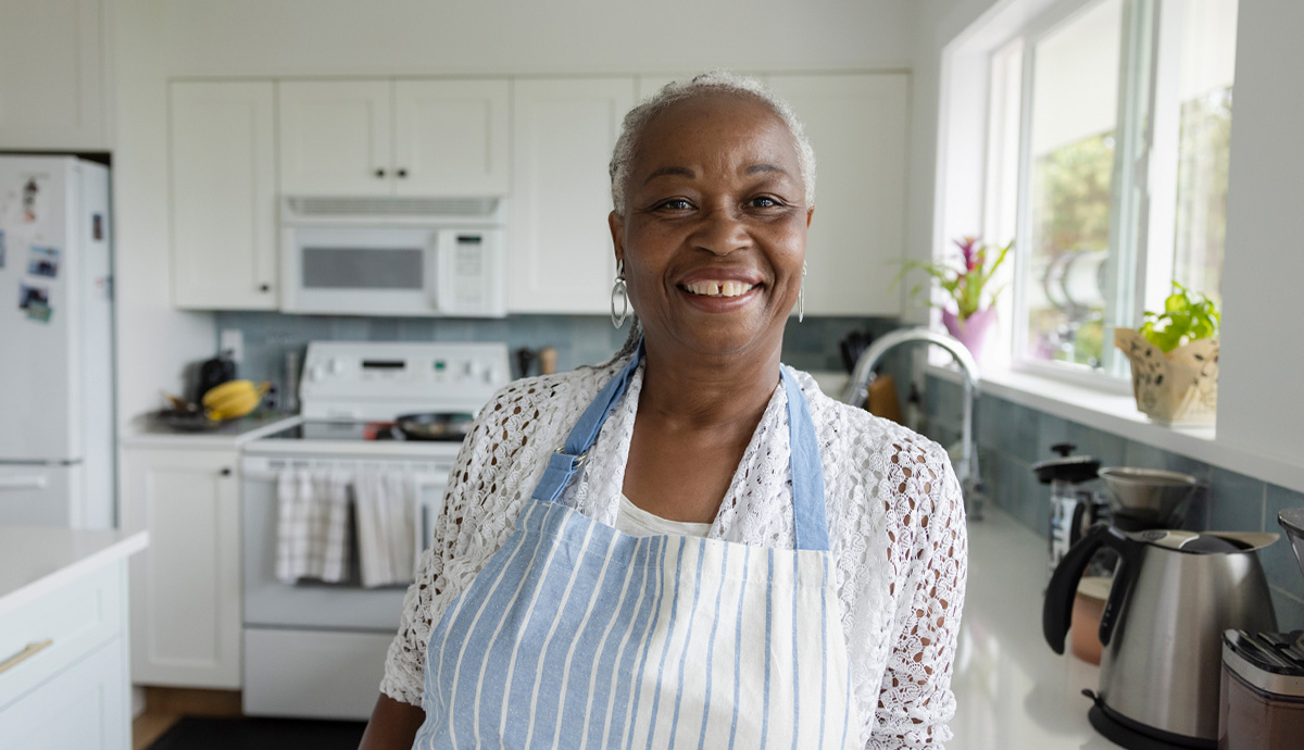 An older woman standing in a kitchen and wearing an apron while smiling at the camera.