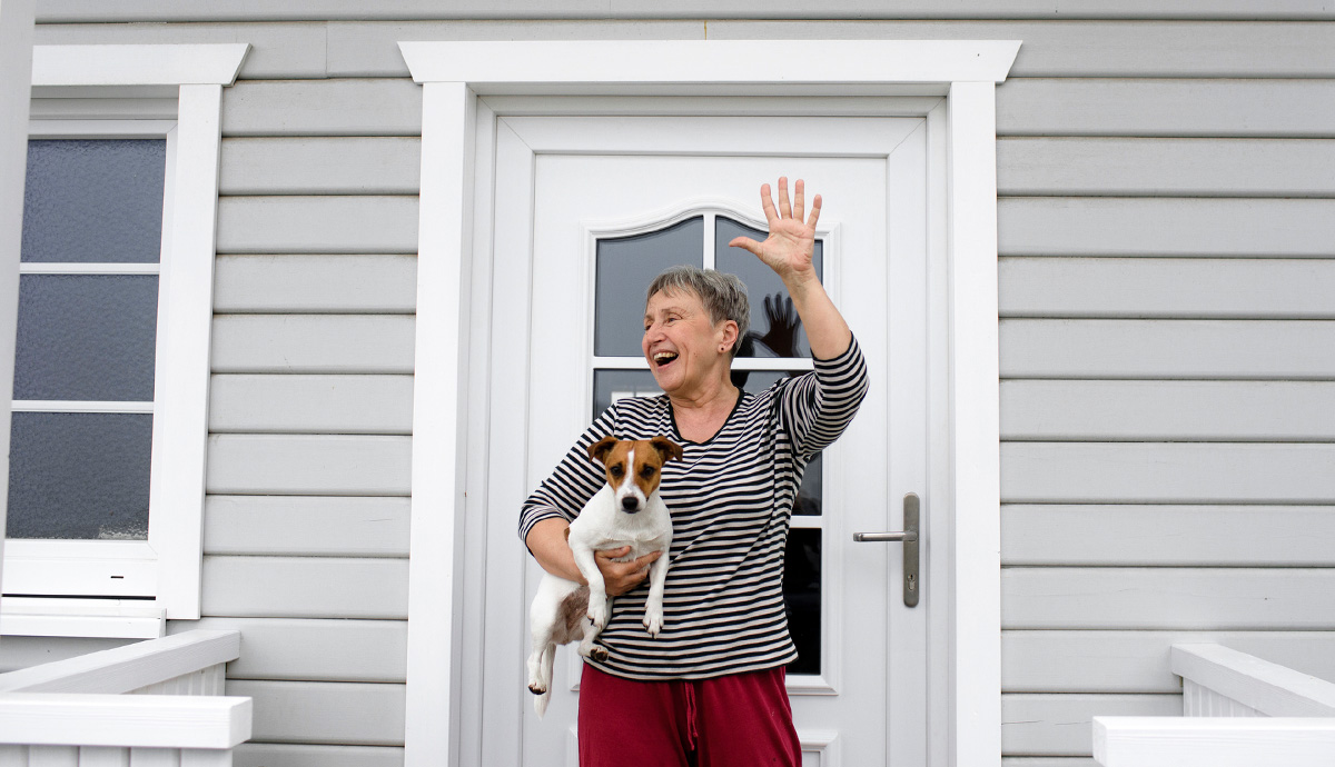 A woman standing in front of her front door holding a small dog in her right arm and waving with her left.