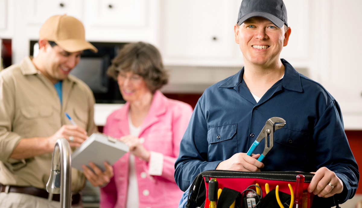 Plumber in uniform stands smiling at the camera with a wrench in hand. Blurred in the background is a man reviewing a clipboard with an older woman.